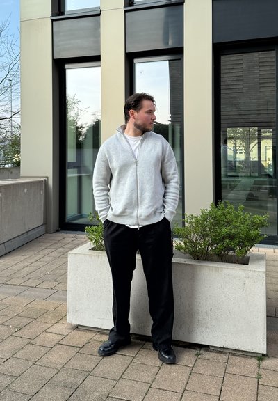 Jersey gris claro con cremallera y cuello alto, usado sobre una camisa blanca; pantalones negros de corte holgado; de pie junto a un macetero de concreto.