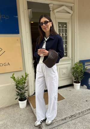 Modelo con una chaqueta corta azul marino, camisa blanca y pantalones anchos blancos, sosteniendo una taza y un bolso de mimbre marrón. Zapatillas grises completan el atuendo.