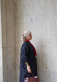 Cappotto lungo di un blu scuro con texture, sciarpa rossa e foulard con motivo. Borsa marrone con dettagli in metallo. Gli accessori includono braccialetti.