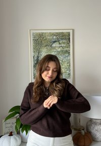 Brązowy sweter prążkowany, białe spodnie z wysokim stanem, z teksturowanymi elementami dekoracyjnymi, takimi jak dynia i pluszowa roślina. W tle sztuka z motywem zieleni.