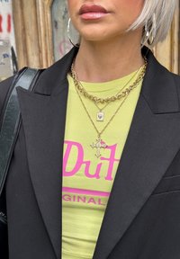 Sort blazer over en grøn t-shirt med pink tekst, prydet med lagdelte guldkæder og et korsvedhæng. Store sølv hoopsøreringe.