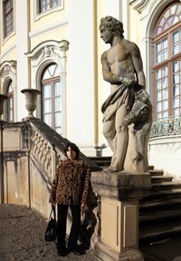 Jeune personne en manteau à imprimé léopard tenant un sac noir se tient près d'une statue en pierre d'un homme tenant une tête d'animal devant un bâtiment classique.