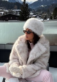 Puhast bež umetnega krzna plašč z izrazito teksturo, kombiniran z ujemajočo krzneno kapo. Model nosi črna sončna očala in svetlo roza obleko.