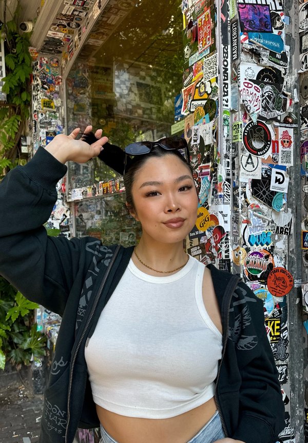 Zwarte zip-up hoodie met grafische accenten over een witte geribbelde crop top. Achtergrond gevuld met kleurrijke stickers op een glazen muur.