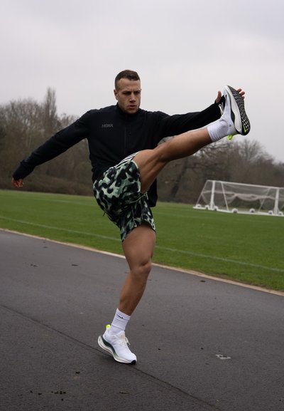 Hombre atlético con chaqueta negra de cremallera y pantalones cortos verdes con un patrón, realizando una patada alta y usando zapatillas de running blancas y verdes.