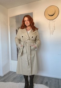 Beige trenchcoat met dubbele rij knopen, een ceintuur op taillehoogte en twee zijzakken, gecombineerd met zwarte kniehoge laarzen op een houten vloer.