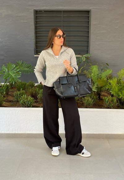 Jersey gris con canalés y media cremallera, pantalones de pata ancha negros y un bolso texturizado negro. Zapatillas blancas con detalles en gris. Plantas verdes de fondo.