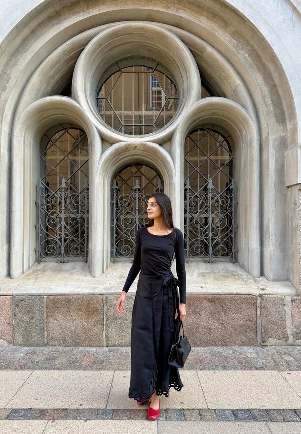 Mujer con top negro y falda larga, con zapatos rojos y bolso, de pie frente a ventanas arqueadas de piedra ornamentadas con rejas de metal.