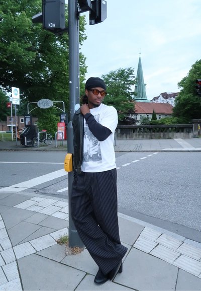 Persona que lleva una chaqueta de cuero negra sobre una camiseta gráfica blanca, pantalones a rayas y zapatos negros, de pie junto a un sem áforo.