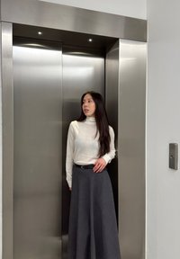 Maglione bianco a collo alto, gonna nera A-line grigio scuro. Porta dell'ascensore con finitura in acciaio inox e luci incassate. Accento con cintura semplice.