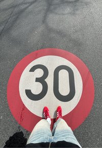 Rdeča okrogla prometna oznaka na pločniku z belo ozadjem, ki prikazuje "30" v poglobljenem črnem tisku. Rdeči teniski čevlji so delno vidni spodaj.