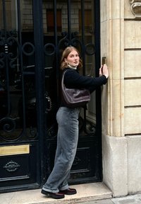 Bruine leren schoudertas, zwarte gebreide trui, witte coltrui, grijze wijde jeans, zwarte platform schoenen, sierlijke zwarte poort met gouden accenten.