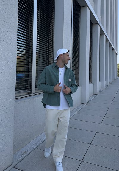 Chaqueta verde clara de botonadura, camiseta blanca, pantalones crema y zapatillas blancas. De pie al aire libre frente a un edificio moderno con grandes ventanales.