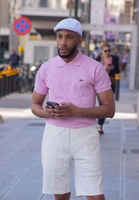 Polo rosa chiaro con maniche corte e un piccolo logo, abbinato a pantaloni corti bianchi fino al ginocchio e un cappellino bianco. In mano ha uno smartphone.