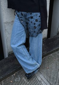 Personne portant un jean large bleu clair, des bottes noires à bout pointu, un pull noir et une jupe superposée noire transparente à motif floral.