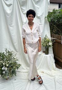 Hvid strikket cardigan med guldknapper over en creme satin slip dress. Båret med sølv sandaler. Blomsterarrangementer i baggrunden.