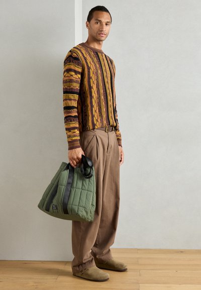 Suéter de punto estampado en tonos tierra, pantalones de pierna ancha en beige y un bolso acolchado verde con correas negras. Look casual con texturas mínimas.