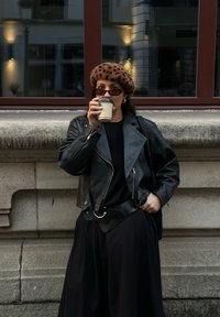 Czarna skórzana kurtka motocyklowa, czarny top, zwiewna czarna spódnica, beret w panterkę oraz okulary przeciwsłoneczne. Trzyma filiżankę kawy.