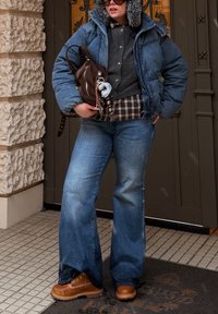 Džínová bunda s kapucí vyloženou kožešinou, šedý svetr, kostkovaná košile, světle modré zvonové džíny a hnědé boty na šněrování. Hnědá kabelka přes rameno s přívěskem.