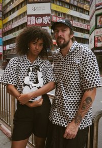 Zwei Personen stehen vor bunten Schildern. Sie tragen passende kurzärmelige Hemden mit schwarzen Herzmustern und schwarze Shorts. Eine von ihnen hält eine schwarz-weiße Tasche.