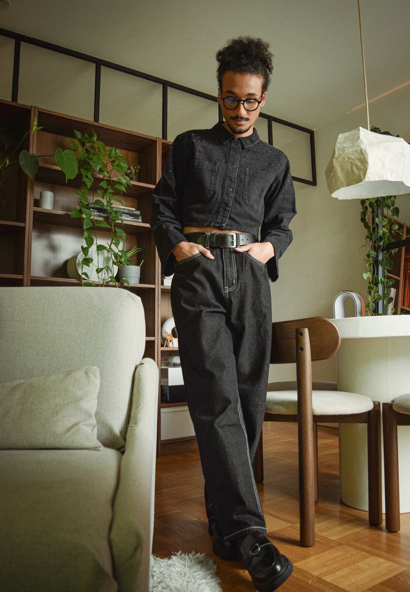 Chemise noire raccourcie avec détails cloutés, jean noir taille haute avec couture blanche, ceinture noire, le tout assorti à des chaussures noires.