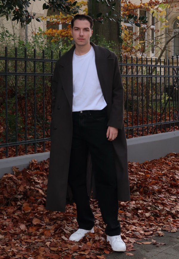 Cappotto lungo verde scuro sopra una maglietta bianca e pantaloni neri, abbinato a sneakers bianche; in piedi su foglie cadute con una recinzione sullo sfondo.