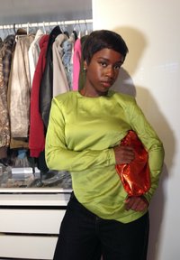 Blusa de mangas longas, verde-lima, com uma textura brilhante e design drapeado, combinada com uma clutch metálica laranja. Jeans escuros usados em baixo.