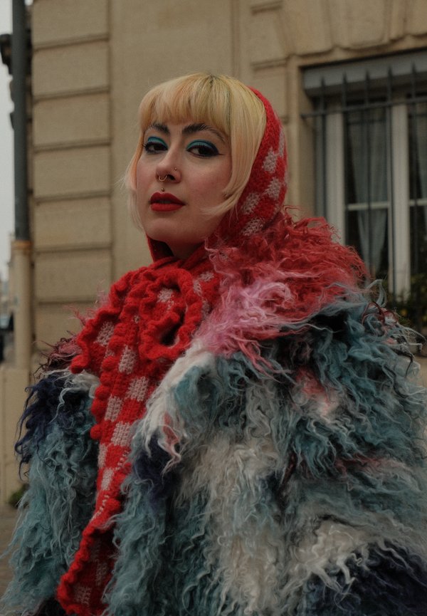 Kleurrijke, pluizige buitenkleding met een mix van blauwe, witte en roze vacht, gecombineerd met een rood geruit gebreide sjaal en capuchon.