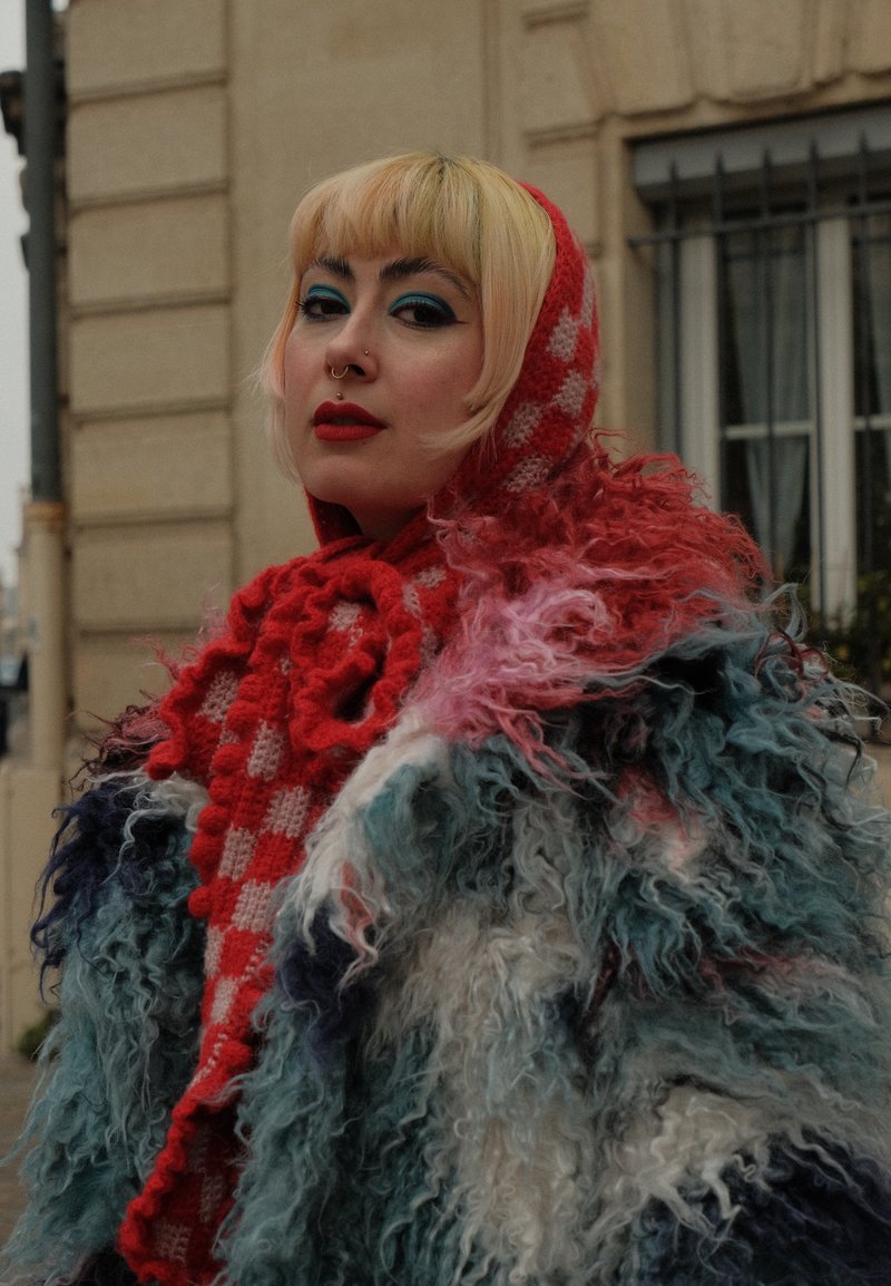 Vêtement d'extérieur coloré et duveteux avec un mélange de fourrure bleue, blanche et rose, associé à une écharpe et une capuche tricotées à carreaux rouges.