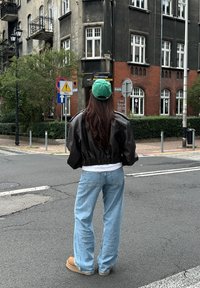 Black cropped leather jacket, white T-shirt, light blue jeans, tan boots, and a green cap. Hair is long and wavy. Urban street setting.