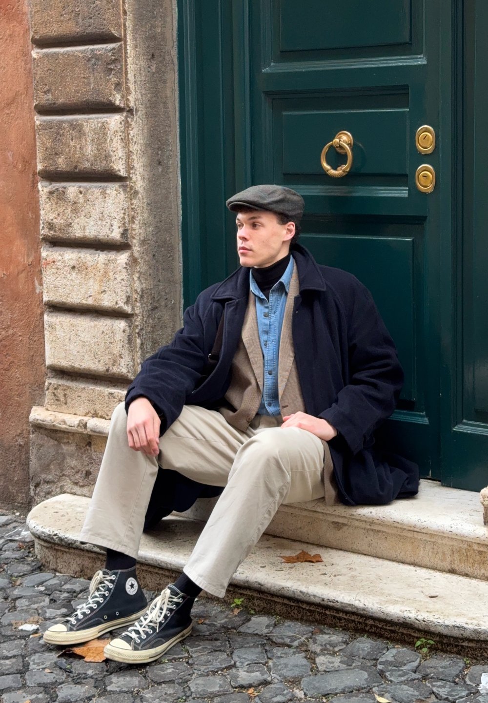 Jeune homme portant des vêtements superposés et une casquette plate, assis sur une marche en pierre devant une porte verte dans une rue pavée.