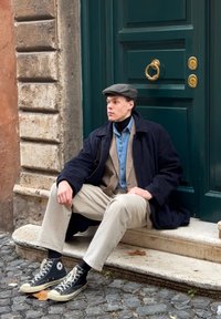 Jeune homme portant des vêtements superposés et une casquette plate, assis sur une marche en pierre devant une porte verte dans une rue pavée.