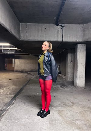 Mujer joven de pie en un estacionamiento de concreto, vestida con una chaqueta de cuero negra, camiseta de rayas y pantalones cortos, medias rojas brillantes y zapatos negros.