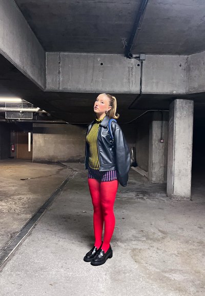 Mujer joven de pie en un estacionamiento de concreto, vestida con una chaqueta de cuero negra, camiseta de rayas y pantalones cortos, medias rojas brillantes y zapatos negros.