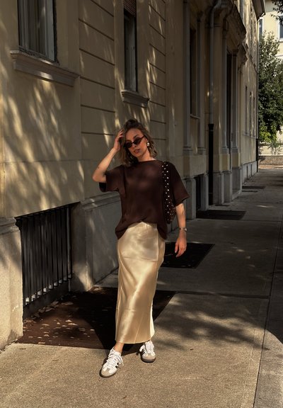 Mujer con gafas de sol, top marrón oscuro, falda de satén beige, zapatillas blancas y bolso de hombro con tachuelas caminando por una acera urbana iluminada por el sol.