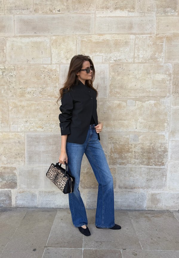 Mujer con chaqueta negra, vaqueros acampanados azules, zapatos negros y gafas de sol sostiene un bolso con estampado de leopardo contra una pared de piedra beige.