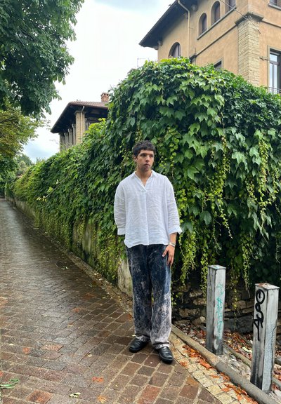 Camisa blanca de lino, pantalones holgados a rayas marinos y gris claro, zapatos negros, de pie junto a un seto verde frondoso en un camino de ladrillos.