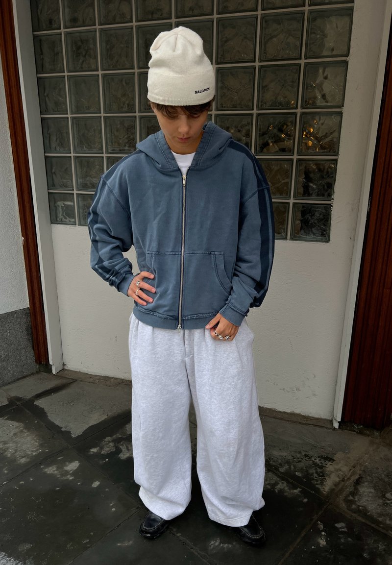 Jeune personne portant un bonnet blanc, un sweat à capuche bleu oversize zippé, un pantalon blanc ample et des chaussures noires, debout à l'intérieur la tête baissée.