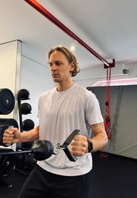 Egy fehér rövid ujjú inges férfi edzést végez egy kétfogantyús csigás rendszer segítségével, amelyen fekete golyós súly van, egy edzőtermi környezetben.