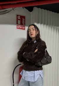 Jeune femme aux longs cheveux foncés, bras croisés, debout près d'un extincteur et d'un panneau rouge « EXTINTOR » sur un mur blanc.