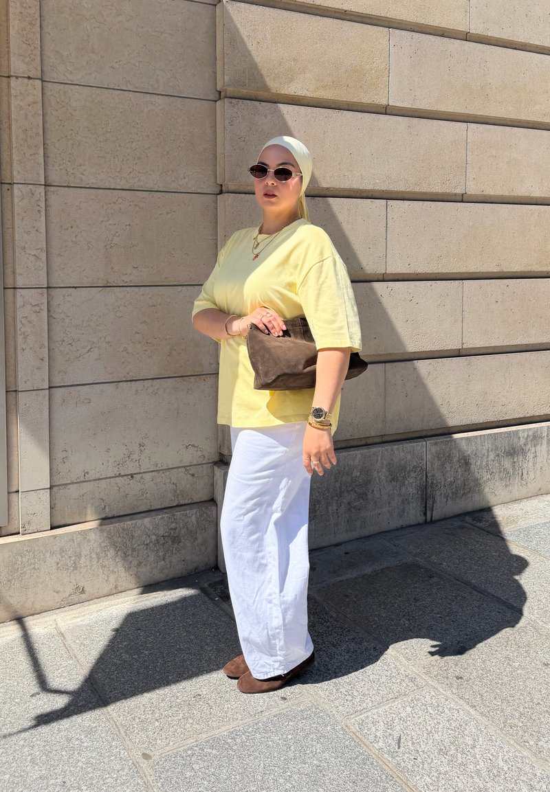 Femme portant une chemise jaune, un pantalon blanc, un foulard sur la tête et des lunettes de soleil, se tient contre un mur de pierre beige, tenant une pochette marron au soleil.