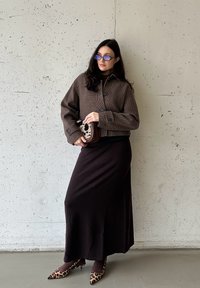 Καφέ καρό κοντό μπουφάν, μαύρο καστόρινο, μακριά μαύρη φούστα και τακούνια με λεοπάρ σχέδιο. Κρατά μια τσάντα με κηλίδες. Λείο σκηνικό από τσιμέντο.