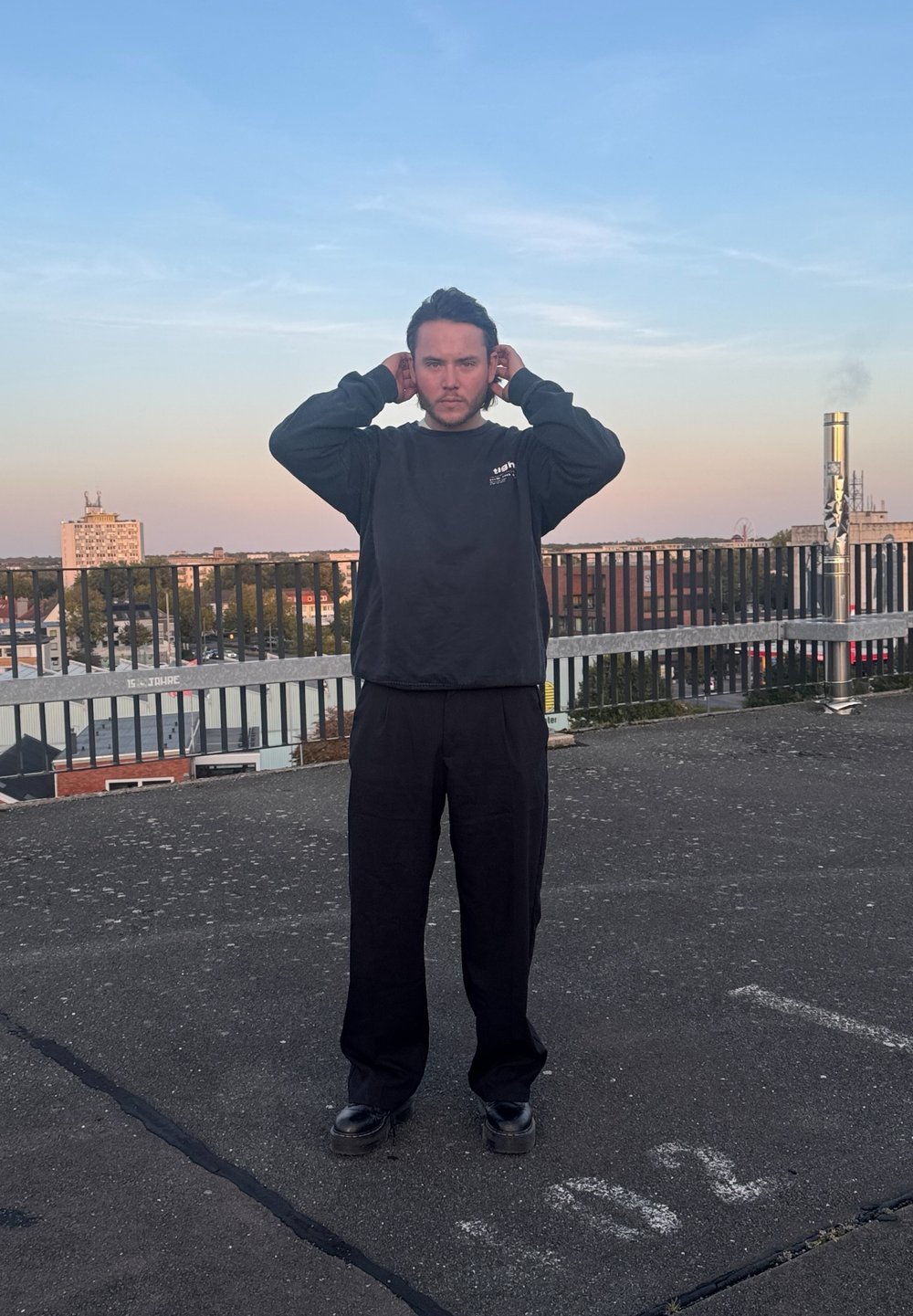 Μαύρη μακρυμάνικη μπλούζα και φαρδιά μαύρα παντελόνια, όρθιοι σε μια ταράτσα με γκρι επιφάνεια και προστατευτική μπάρα στο φόντο.