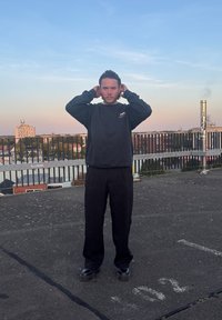 Μαύρη μακρυμάνικη μπλούζα και φαρδιά μαύρα παντελόνια, όρθιοι σε μια ταράτσα με γκρι επιφάνεια και προστατευτική μπάρα στο φόντο.