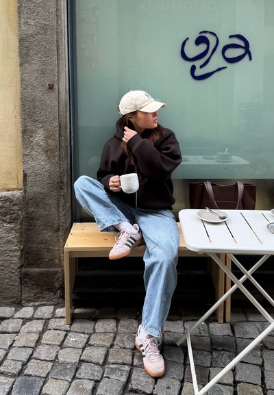 Mujer joven con ropa casual sentada en un banco de madera frente a una cafetería, sosteniendo una taza, mirando de lado cerca de una calle empedrada.