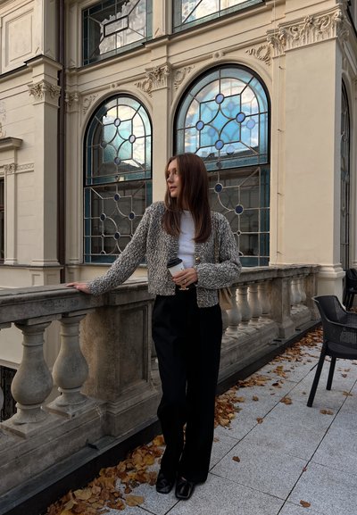 Chaqueta recortada texturizada gris sobre una blusa blanca, pantalones de pierna ancha negros, de pie junto a un balcón de piedra con ventanas de vidrio azul en el fondo.