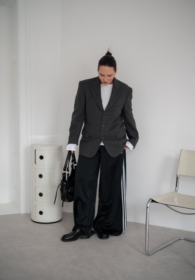 Persona con una americana oscura de gran tamaño, camisa blanca y pantalones negros anchos con rayas blancas, de pie en el interior sosteniendo un bolso negro junto a una silla blanca.