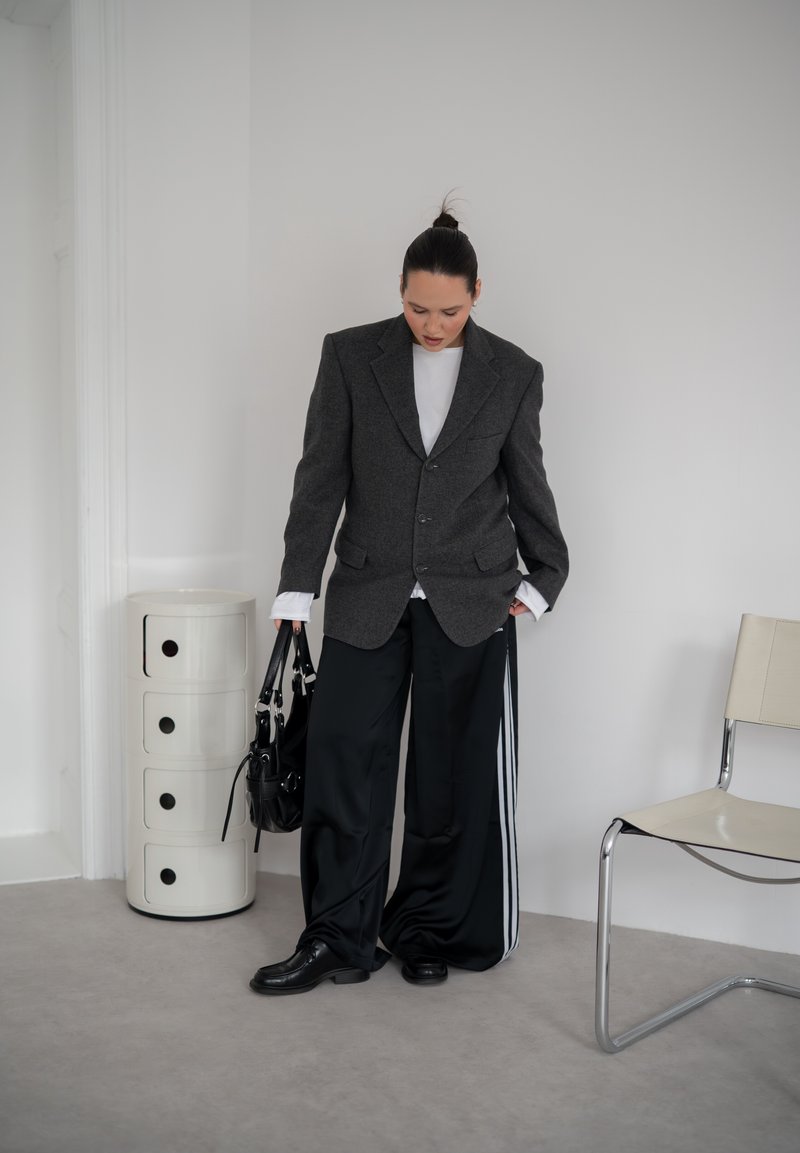 Osoba v nadměrně velkém tmavém blejzru, bílé košili a širokých černých kalhotách s bílými pruhy stojí uvnitř a drží černou kabelku vedle bílé židle.