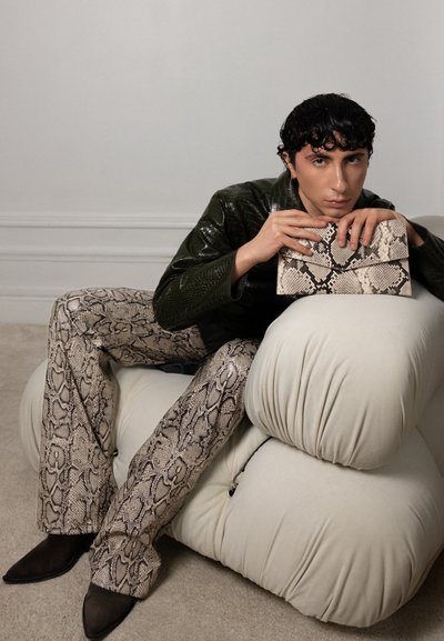 Persona con cabello rizado corto y negro sentada en una silla beige acolchada, vistiendo pantalones con estampado de serpiente y sosteniendo un bolso de mano a juego con el mismo estampado.