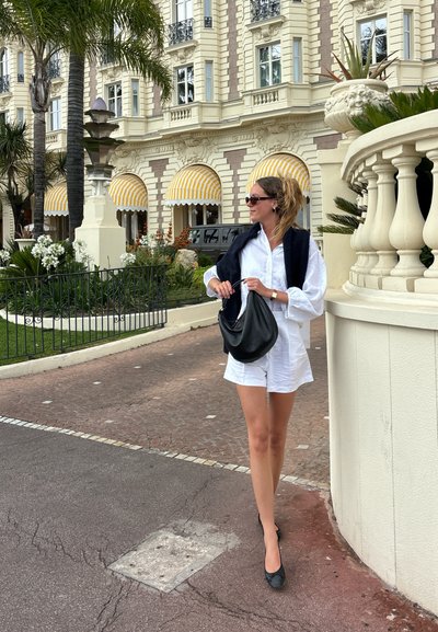 Vestido camisero blanco de lino, bolso negro de hombro, bailarinas negras. El fondo presenta edificios decorativos y palmeras. Toldos con patrones arriba.